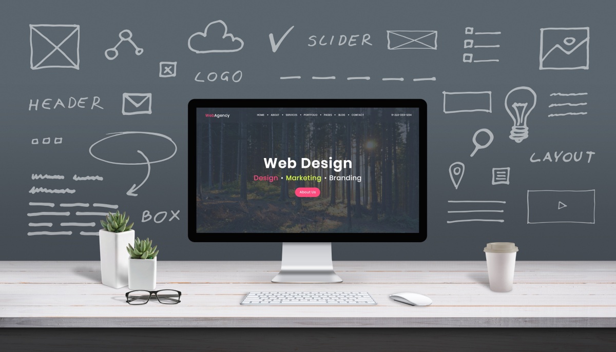 A computer displaying a web design agency website in front of a chalkboard filled with design notes and icons, on a stylish desk.