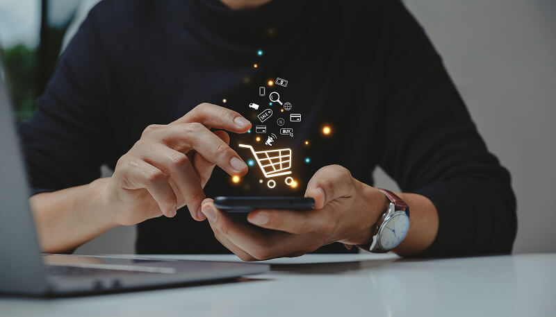 A person uses a smartphone with floating shopping icons like a cart and lock, on a desk with a laptop, illustrating online shopping innovation.
