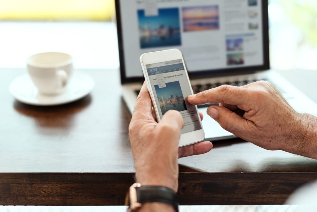 Person reading a news article on a smartphone with a laptop and coffee cup on a desk.