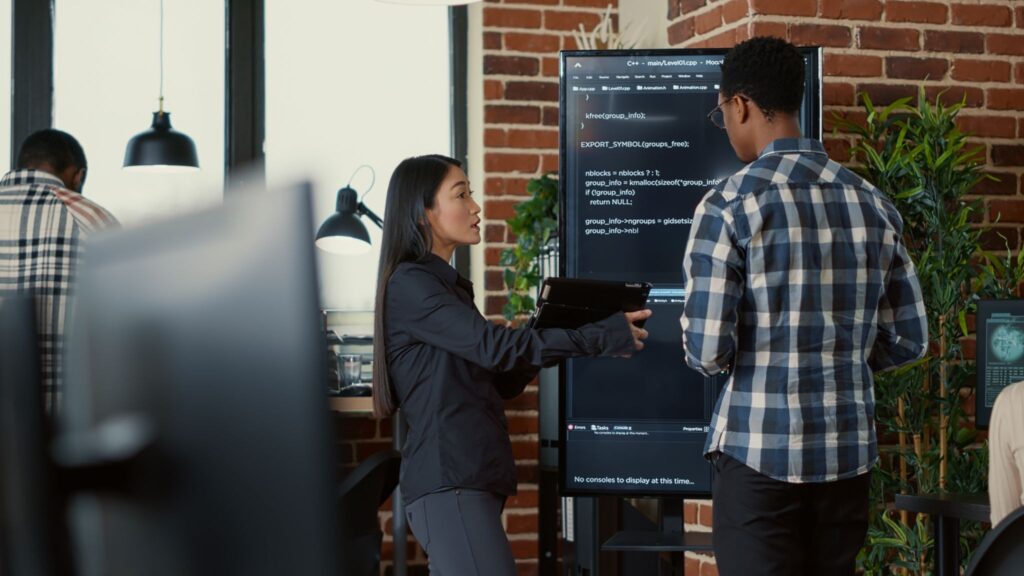 Two developers discussing C++ code displayed on a large wall-mounted screen in an office