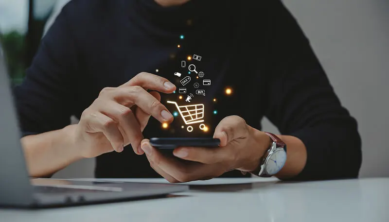 A person uses a smartphone with floating shopping icons like a cart and lock, on a desk with a laptop, illustrating online shopping innovation.